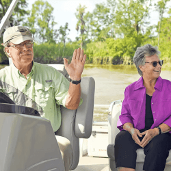 An older man waves from the helm of a boat while an older woman smiles beside him on the water